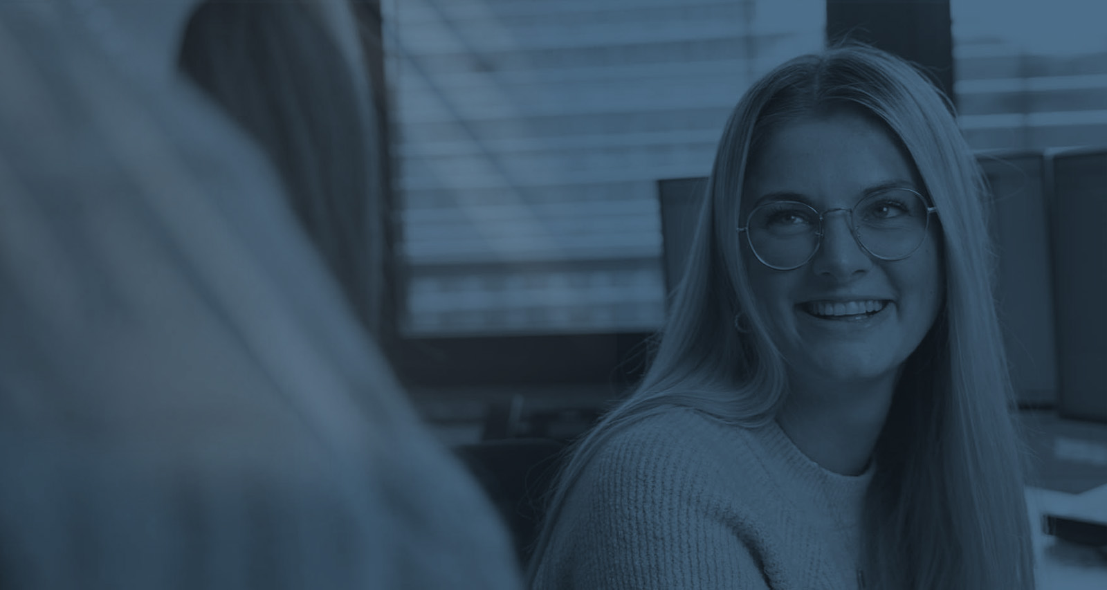 Eine junge Frau mit Brille lächelt in einem Büro und spricht mit einer anderen Person.